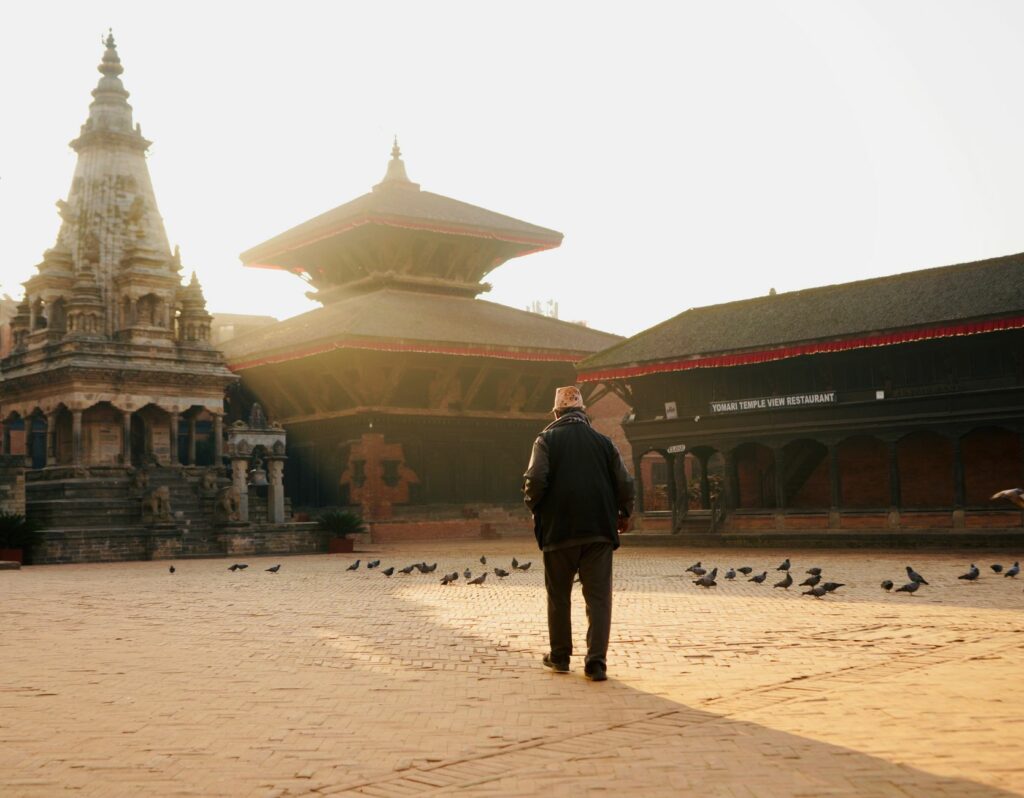 Historic square in Bhaktapur, Nepal, with traditional brick architecture, temples, pigeons, and a pedestrian.